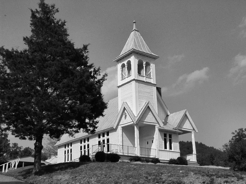 Black and white photograph of a white clapboard country church with a bell tower, standing on a rise beside a large tree